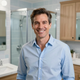 Smiling man in a light blue shirt standing in a bright bathroom with a glass shower and wooden cabinets