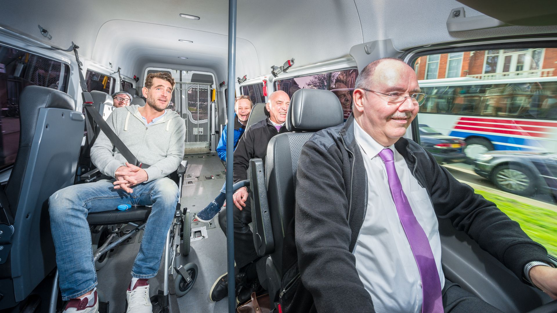 A group of people travelling in a taxi with the driver and a person in a wheelchair upfront. A group of people travelling in a taxi with the driver and a person in a wheelchair upfront.