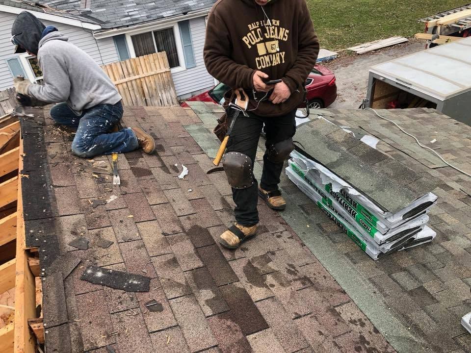 Two roofers repairing shingles on a house roof, one kneeling and one holding tools near stacked materials.