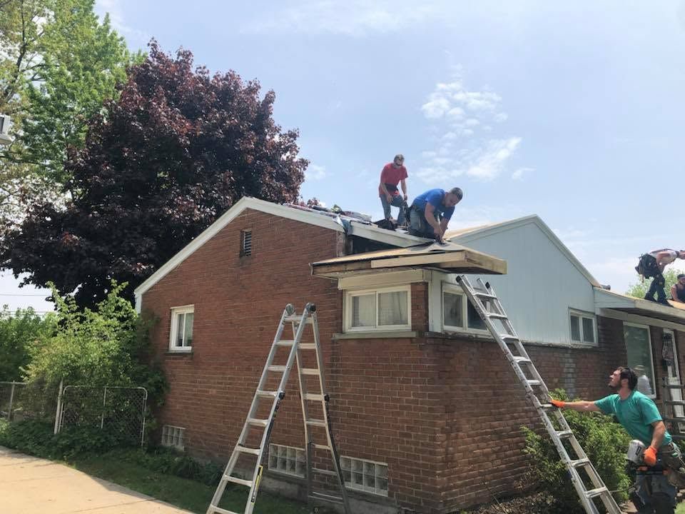 Workers on a roof repairing a brick house, with ladders leaning against the building.