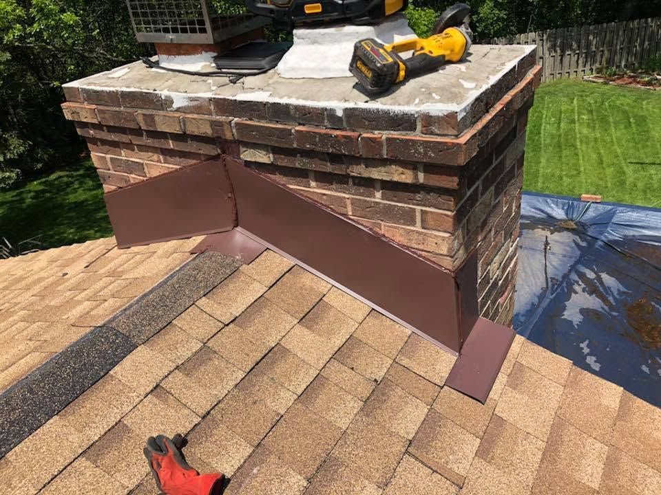 Roof repair tools on a shingled roof beside a brick chimney in a yard.