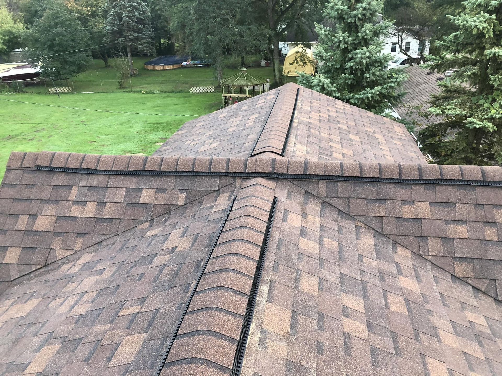 View of a shingled roof with ridgeline, overlooking a green backyard and trees