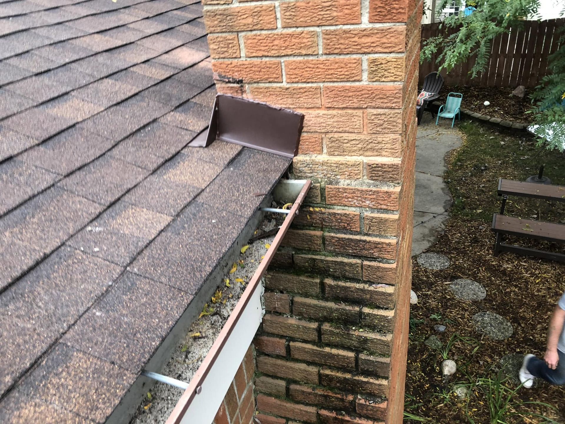Rain gutter and roof shingles beside a brick wall, with a person’s legs visible below in the yard.