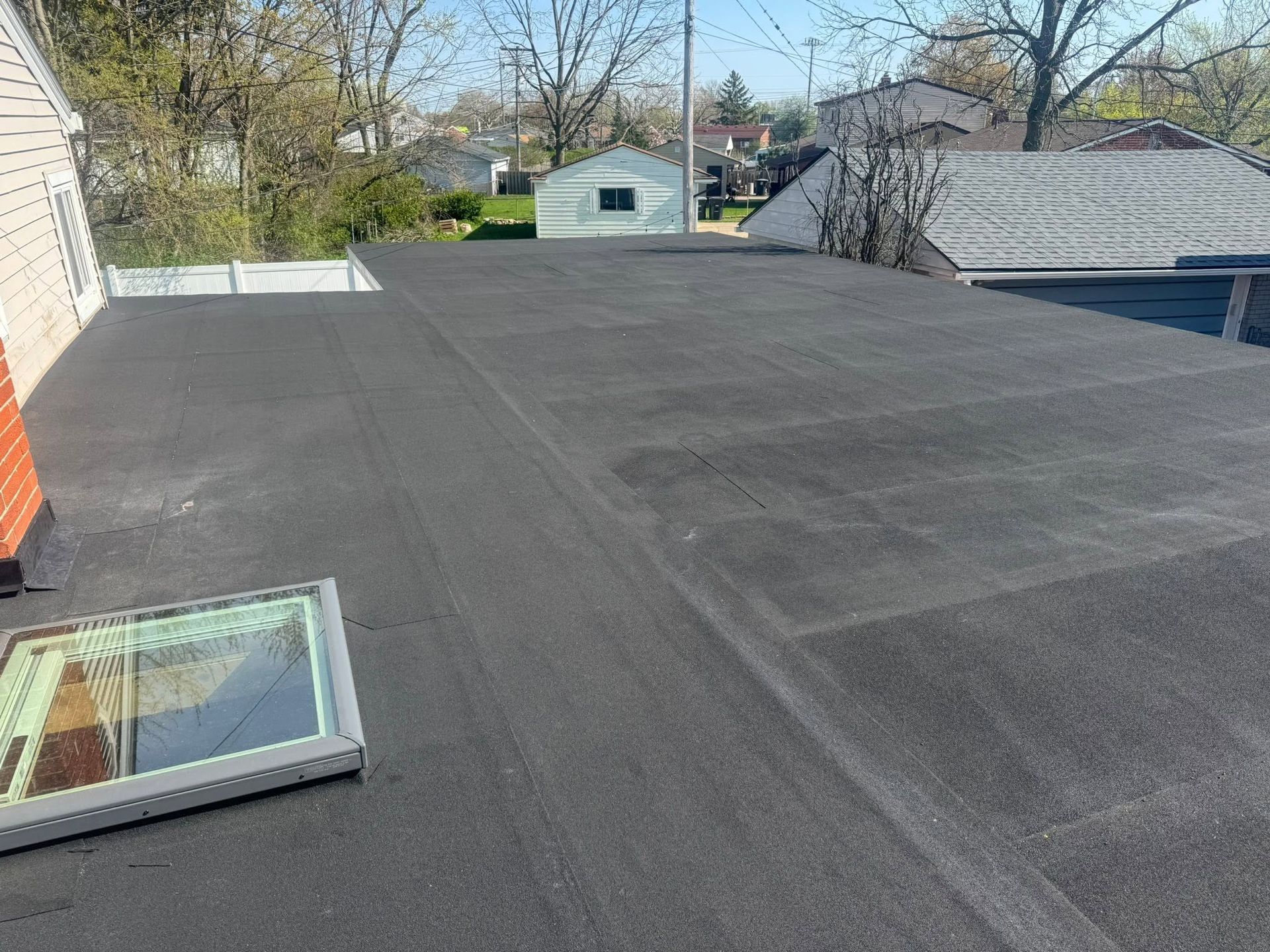 Flat black rooftop with a skylight, trees and neighboring houses visible beyond.
