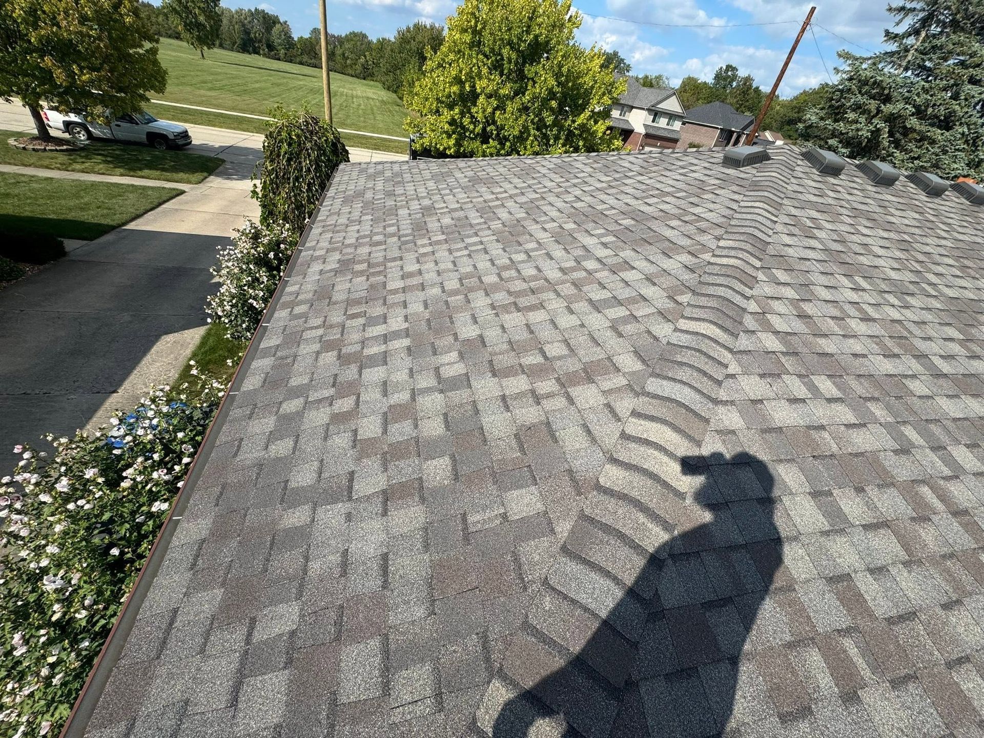 Shingled roof with a person’s shadow, overlooking a driveway and trees on a sunny day