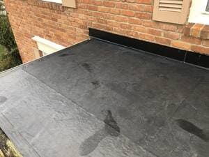 Black flat roof against a brick house wall with a small vent and dark stain marks