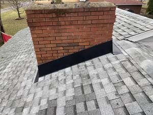 Brick chimney on a gray shingled roof with a black flashing strip at the base.