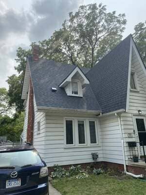 White house with steep gray roof, dormer window, and parked black car in front under cloudy sky