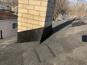 Rooftop with a brick chimney and black flashing, seen beside neighboring houses and leafless trees.