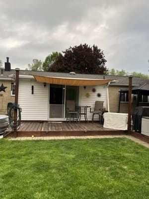 Small backyard deck with white siding, striped awning, two chairs, and green lawn under cloudy sky