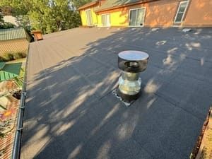 Small silver roof vent on a dark shingled roof in sunlight with tree shadows.