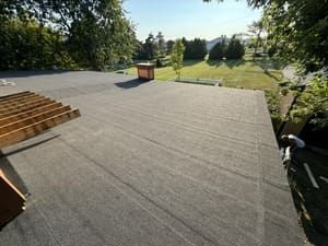 Flat roof under construction with rolled roofing, roof vents, and trees in the background