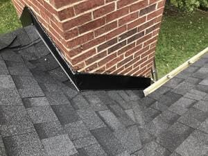 Shingled roof valley around a brick chimney with black flashing and a small metal edging strip