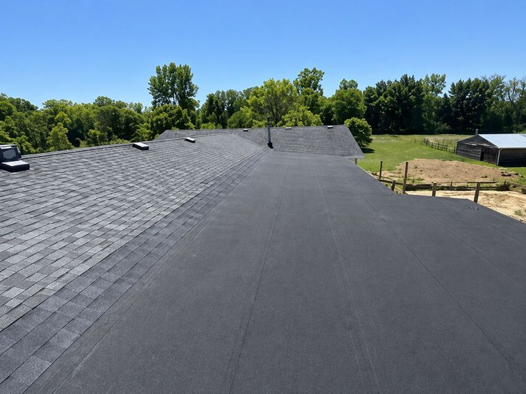 Freshly shingled roof with a long dark ridge under a clear sky, trees and fields in the background Flat black roof under construction beside a green lawn and trees under a clear sky