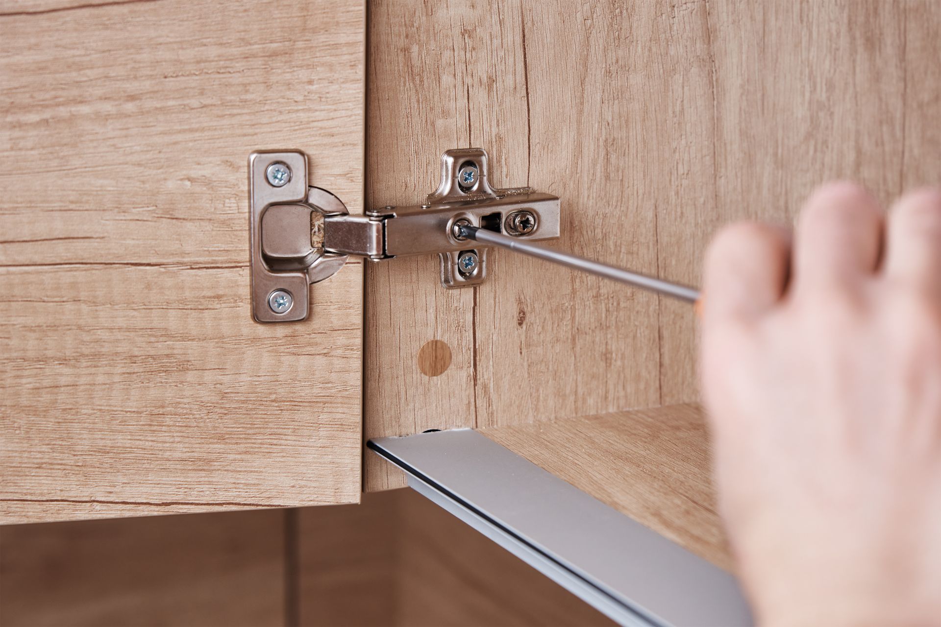 Hand tightening a cabinet hinge with a screwdriver inside a wooden cabinet. Hand using a screwdriver to adjust a metal cabinet hinge on a wooden cabinet door