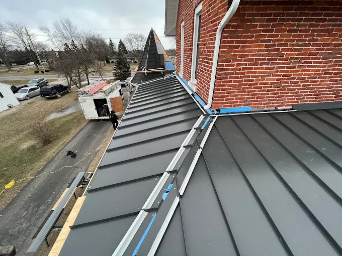 Two power drills on a corrugated metal roof beside scattered screws and a partially attached panel. Two clamps on a corrugated metal roof with rows of fasteners and a strip of greenery at the edge