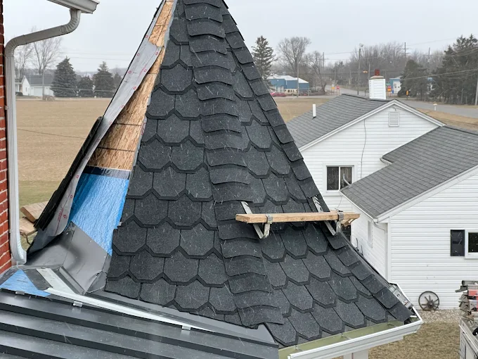 Close-up of a dark gray shingled roof with a roof ridge and suburban houses in the background. Close-up of a new gray shingle roof with gutter and neighborhood houses in the background