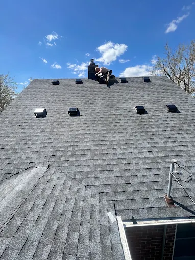 Workers installing gray roof shingles on a steep roof, with stacks of shingles and tools visible. Workers installing dark roof shingles on a sloped roof with tools and stacks of materials.