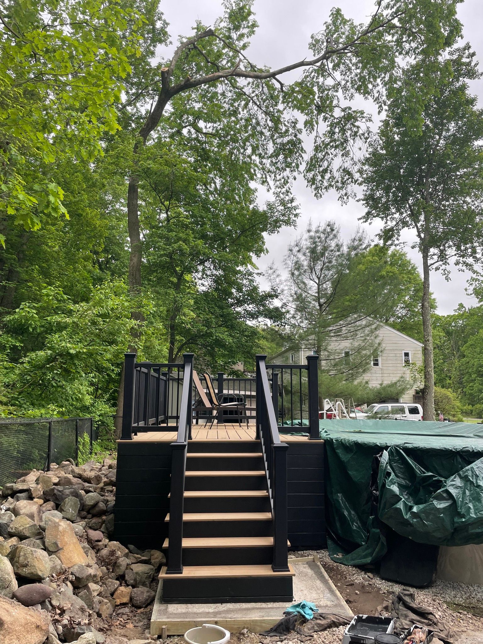 Wooden deck with steps, black railing, surrounded by trees and a tarp-covered pool.