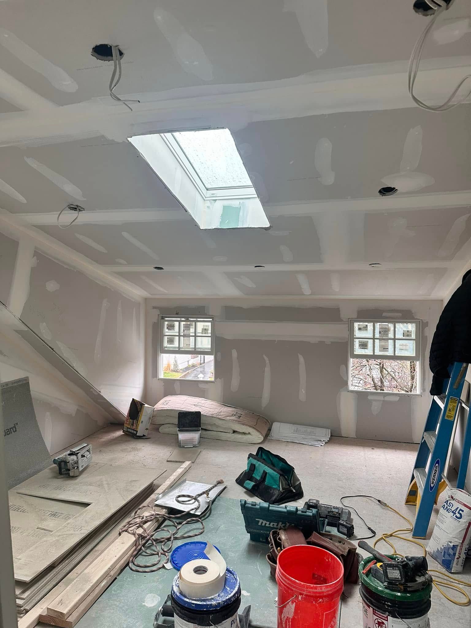 Room under construction with drywall installed on walls and ceiling, skylight, tools and materials scattered.