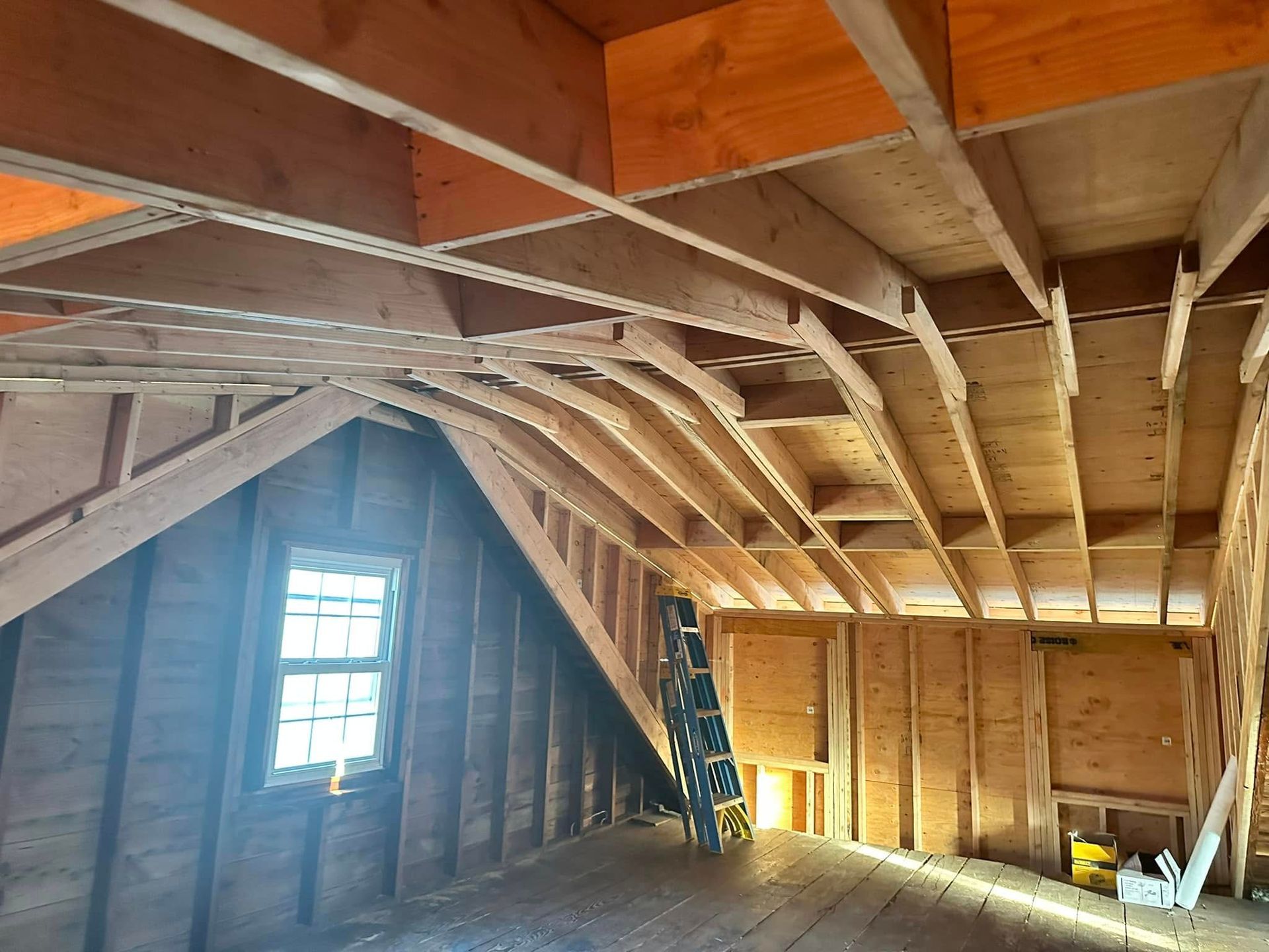 Interior attic with exposed beams and unfinished walls. A ladder stands against a wall, sunlight streams through a window.