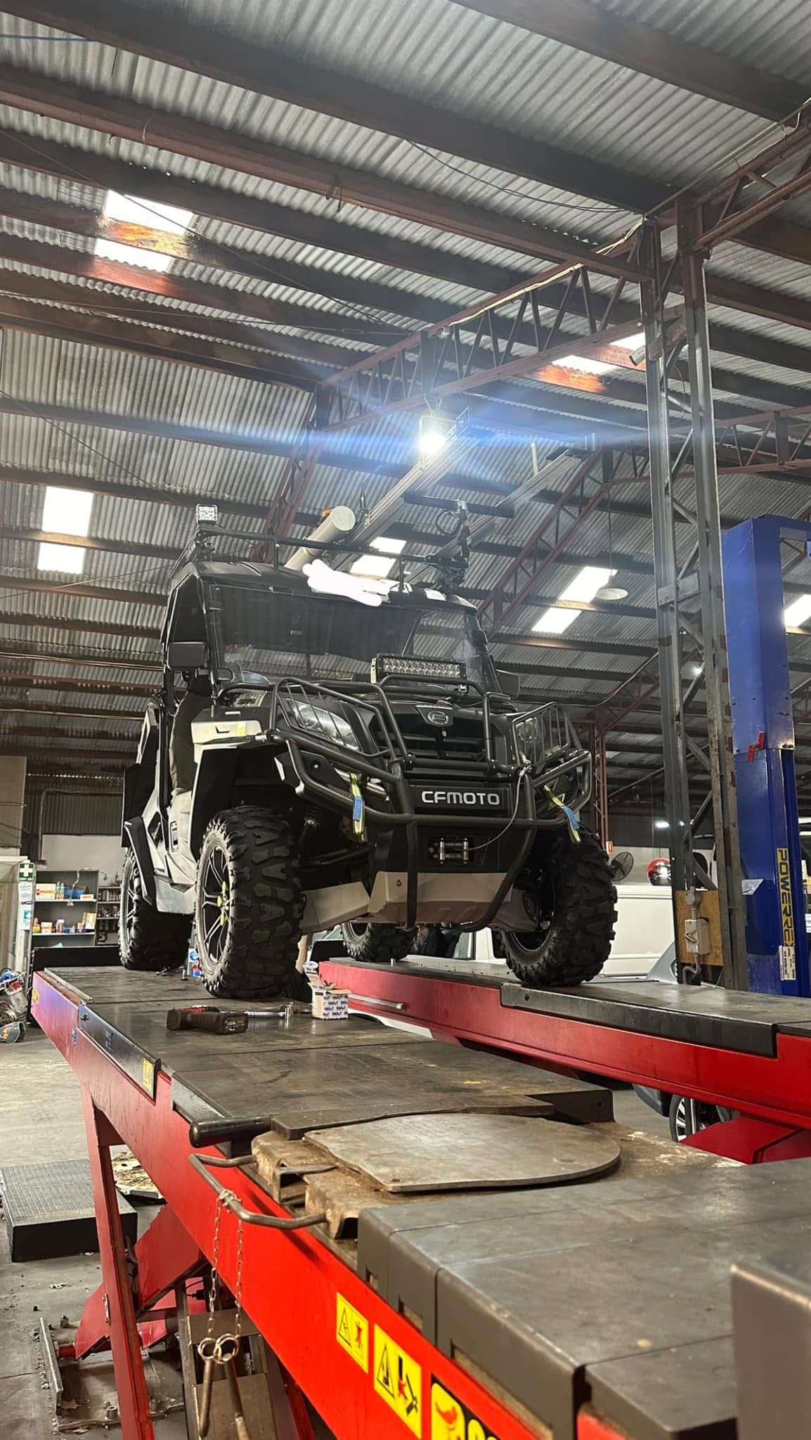 Heavy off-road truck on a lift in a metal-roofed workshop, with red ramps and bright overhead lights. Heavy off-road truck on a lift in a metal-roofed workshop, with red ramps and bright overhead lights.