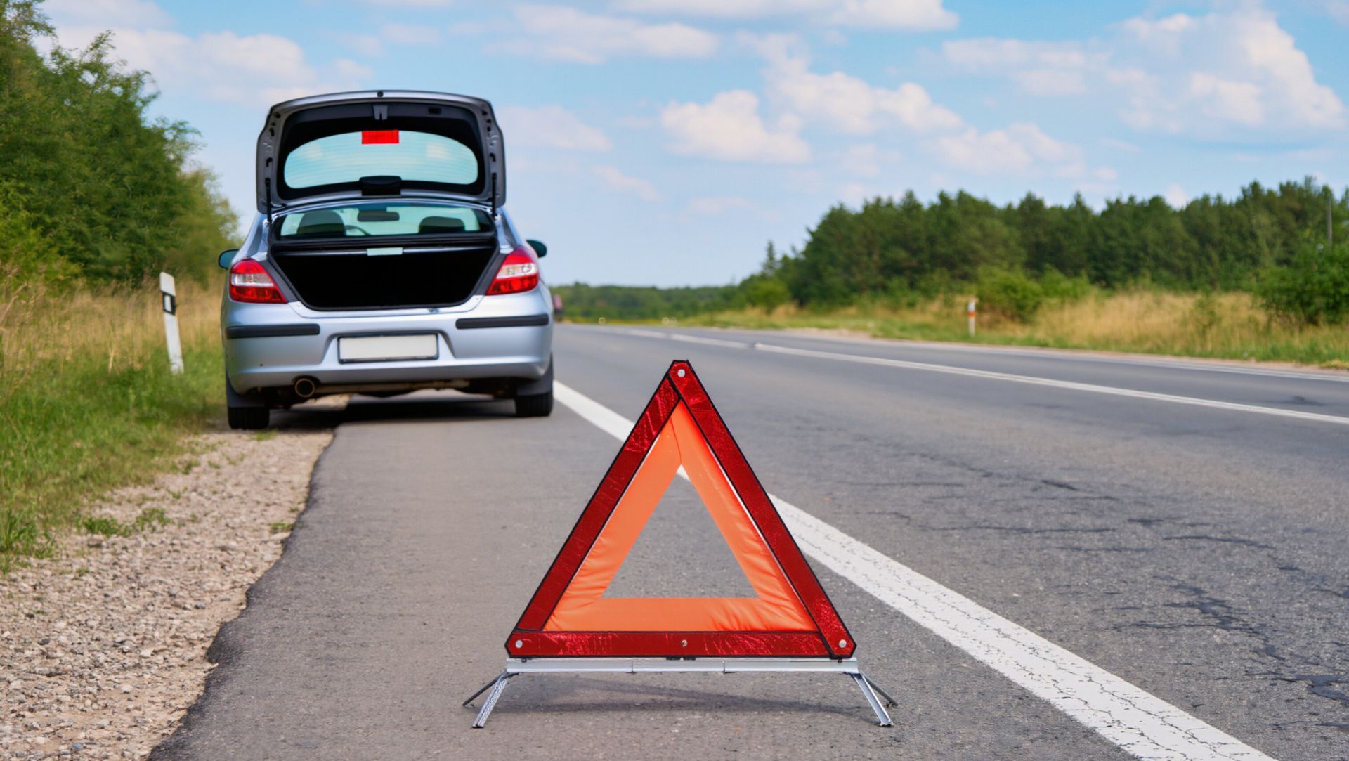 Red warning triangle on roadside behind a stopped silver car with open trunk.