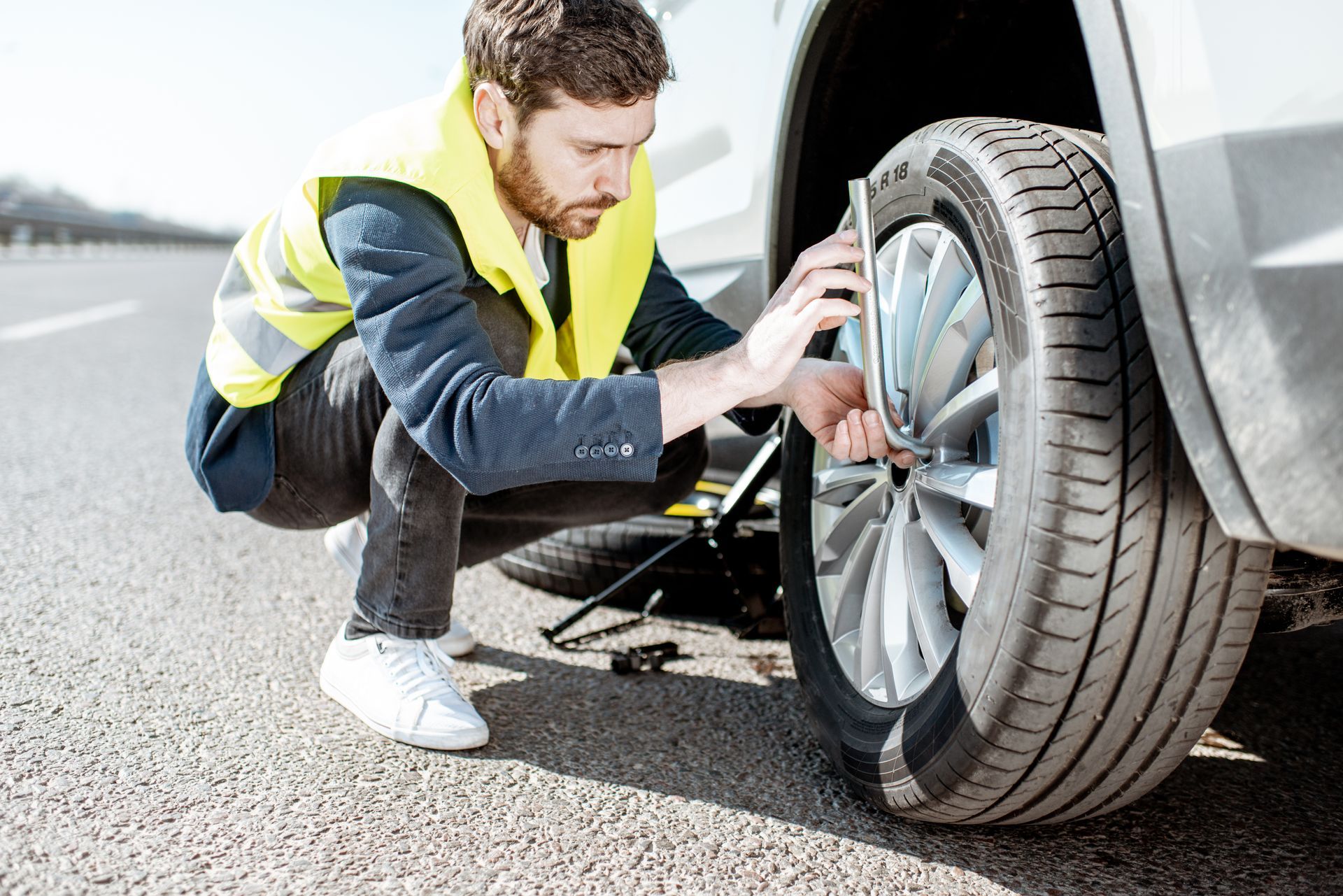 Man kneeling beside a car, checking the front tire with a jack on the roadside.