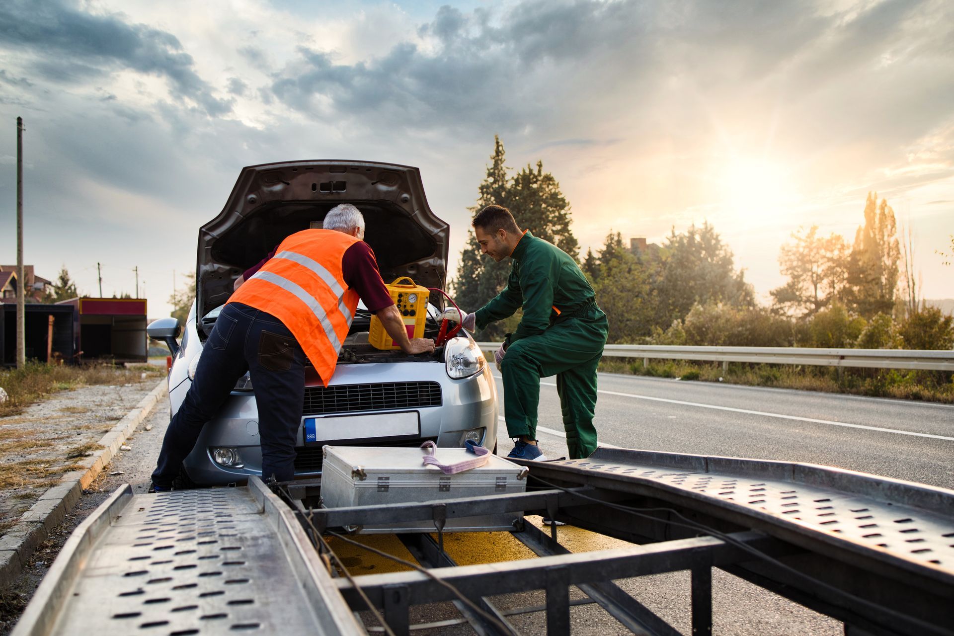 Two workers in safety gear operate equipment on a roadwork platform at sunrise.