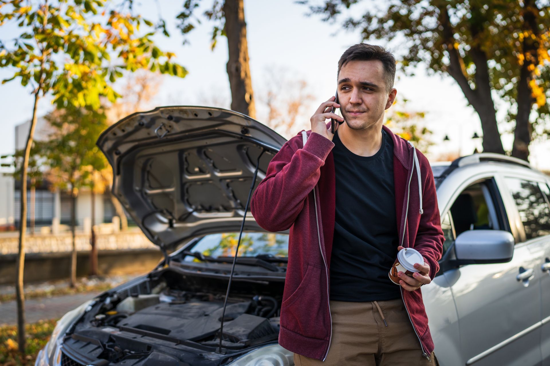 Man standing by a car with its hood open, talking on a phone outdoors, holding a cup