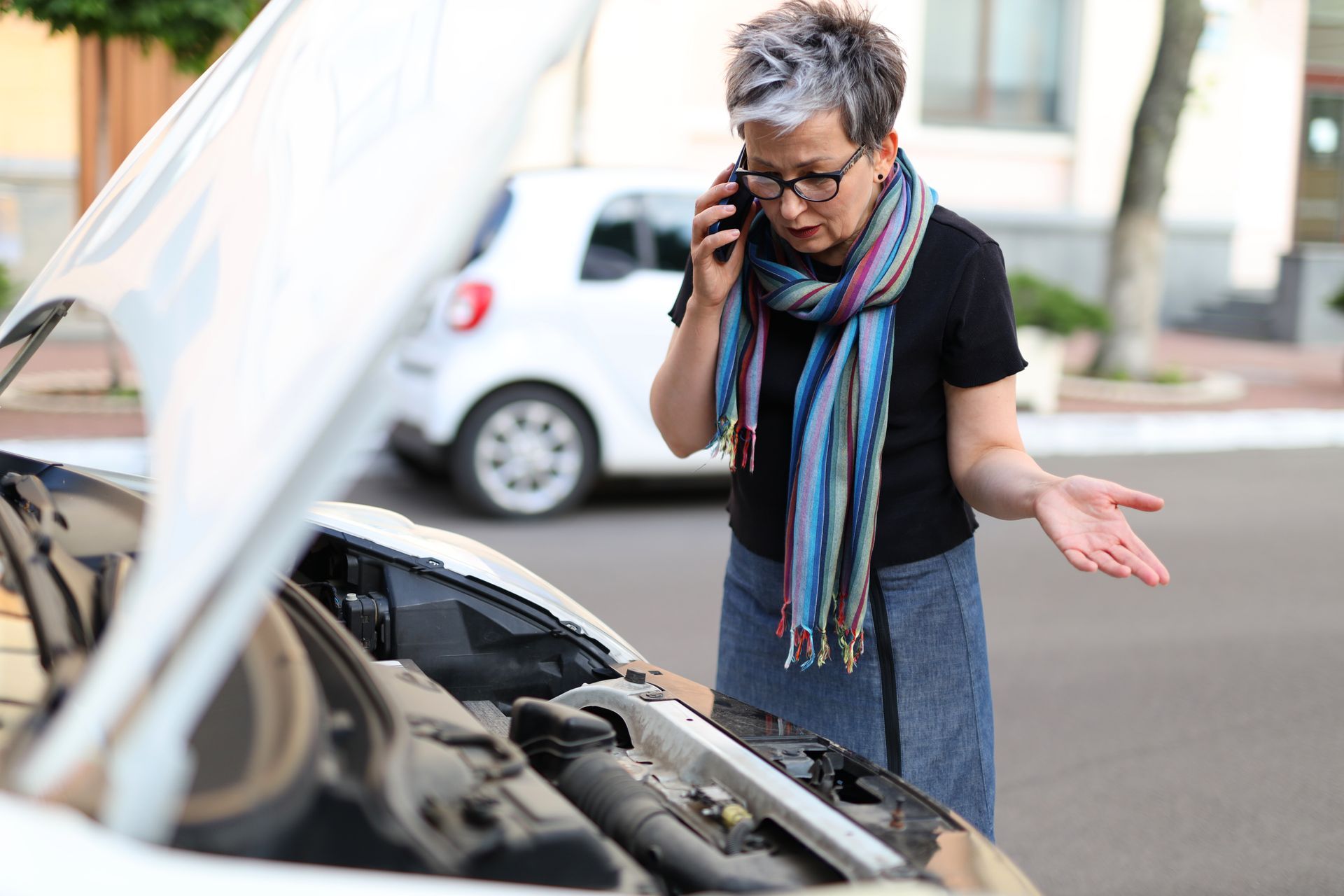 Person on phone beside open car hood, looking frustrated on a street