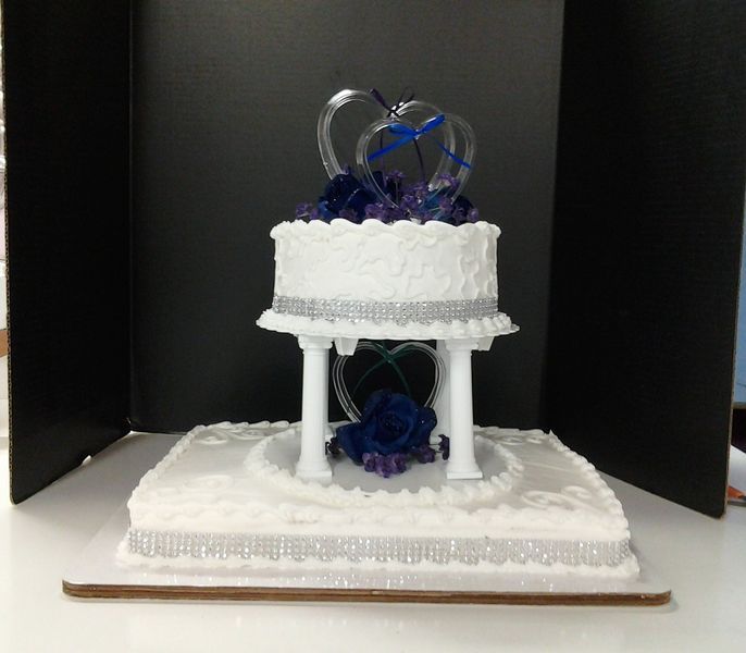 Two-tiered white wedding cake with blue and silver decorations. Clear heart topper.