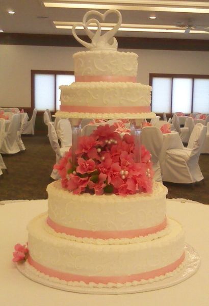 Five-tiered wedding cake with pink ribbon and flowers, topped with swan figures, displayed in a ballroom.
