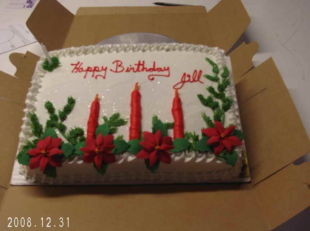 Birthday cake decorated with red candles, poinsettias, and green leaves, in a cardboard box.