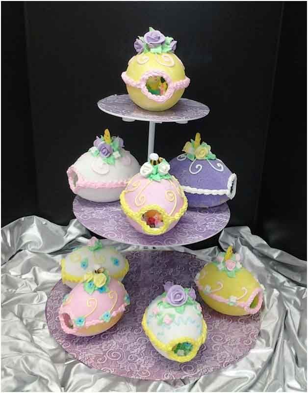 Tiered cake stand with pastel-colored egg-shaped cakes decorated with frosting, flowers, and cut-out windows.