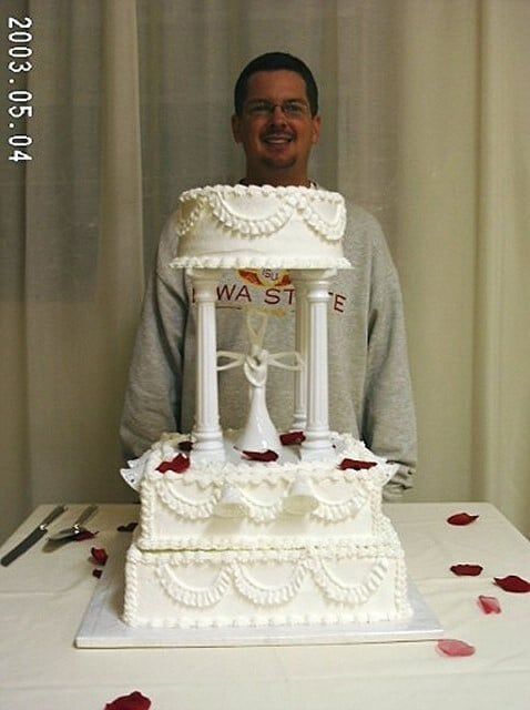 Man smiles behind a multi-tiered wedding cake with white frosting and a knotted decoration; red rose petals on table.