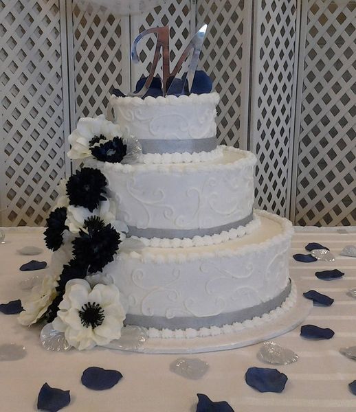 Three-tiered white cake with silver ribbon and dark flowers, topped with the letter 