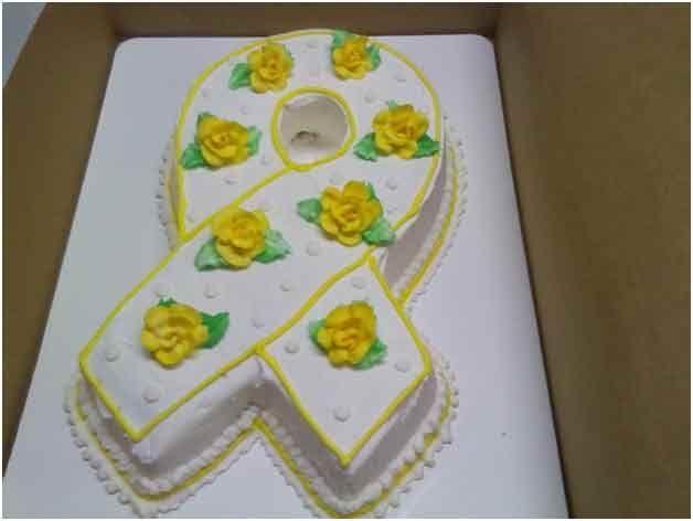 Cake shaped like a yellow ribbon decorated with yellow roses and white frosting, in a box.