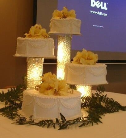 Tiered wedding cake with yellow flower toppers, set on illuminated glass pillars, on a table with greenery.