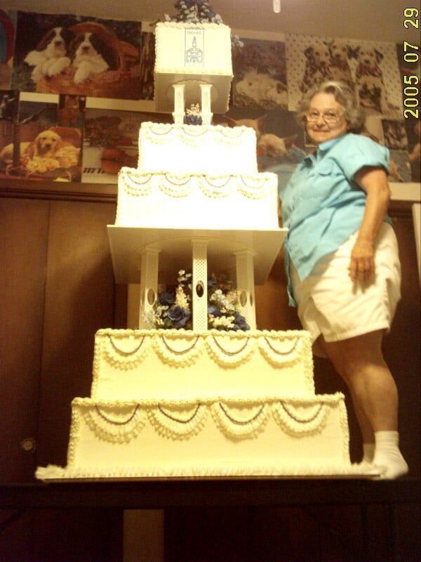Woman stands next to a five-tiered, ornate white cake with blue accents. The date 2005 07 29 is visible.