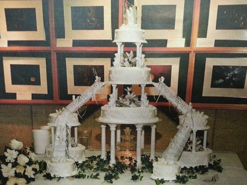 Elaborate white tiered wedding cake with connecting bridges, against a wall of framed art.