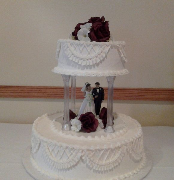 Two-tiered white wedding cake with a bride, groom figurine, and burgundy and white flower decorations.