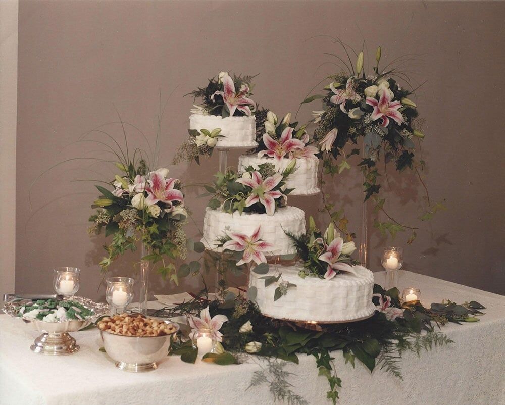 Tiered white wedding cake decorated with lilies and greenery, with floral arrangements and candles.