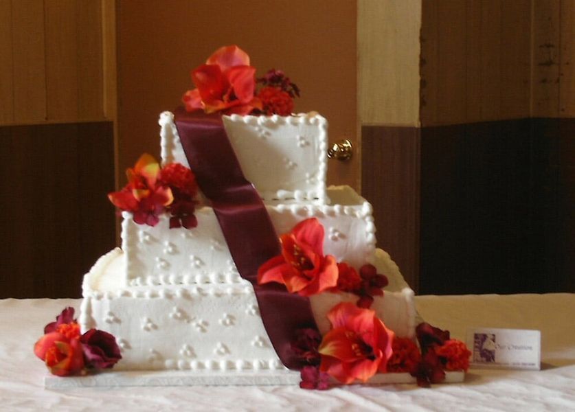 Three-tiered white square cake with maroon ribbon, red flowers, and pearl accents.