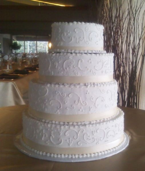 Four-tiered white wedding cake with intricate scrollwork, pearl border, and cream-colored ribbon on a table.