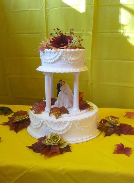 Two-tiered wedding cake with bride and groom figurines, decorated with fall-themed foliage and on a yellow tablecloth.