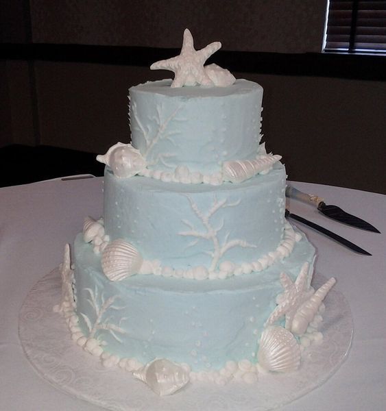 Three-tiered blue cake decorated with white seashells and starfish, on a white tablecloth.