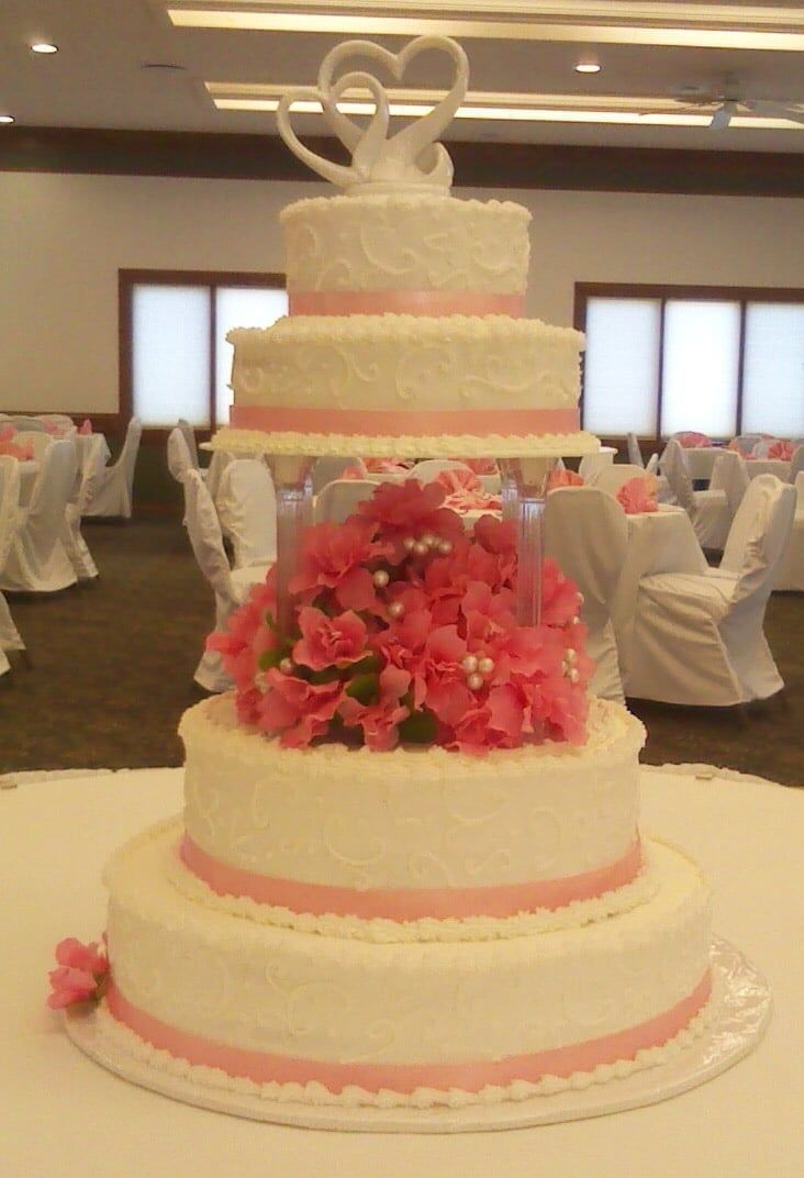 Four-tiered white wedding cake with pink ribbon, flowers, and swan topper, on a table.