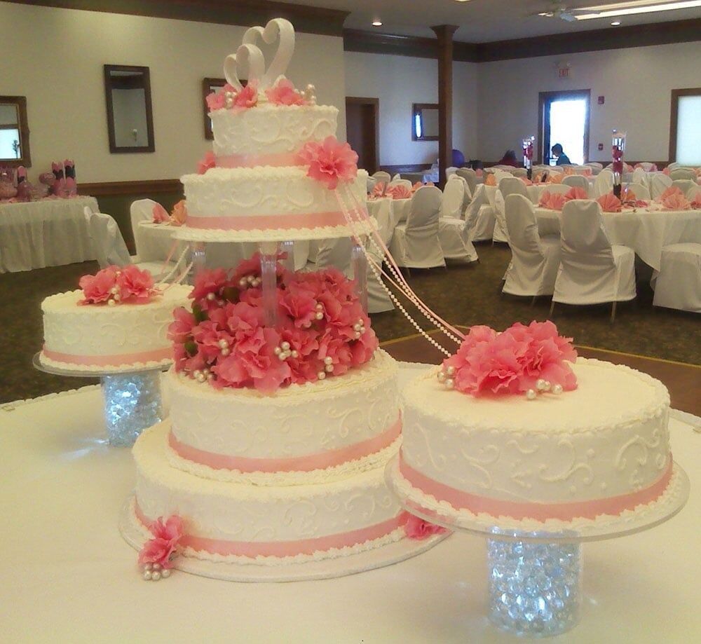 Wedding cakes, tiered and decorated with pink flowers, on a table. Reception hall in background.