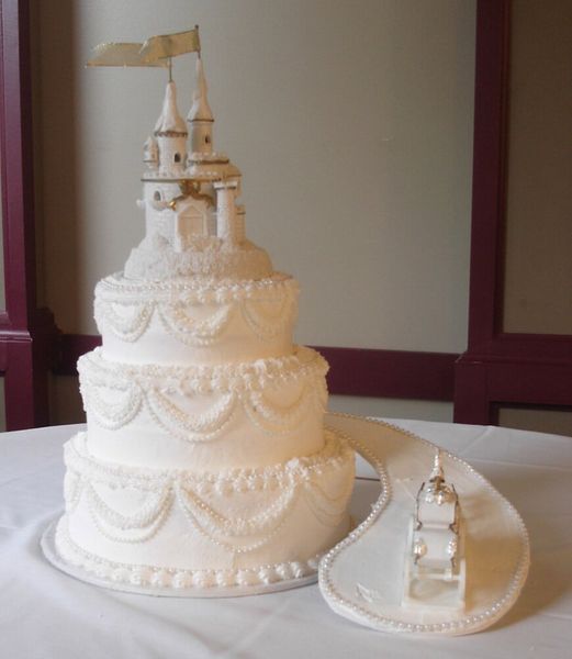 White tiered cake with castle topper and sugar road, on a white tablecloth.